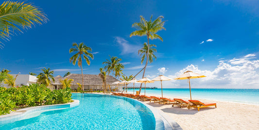 Luxurious beach resort with swimming pool and beach chairs or loungers under umbrellas with palm trees and blue sky.